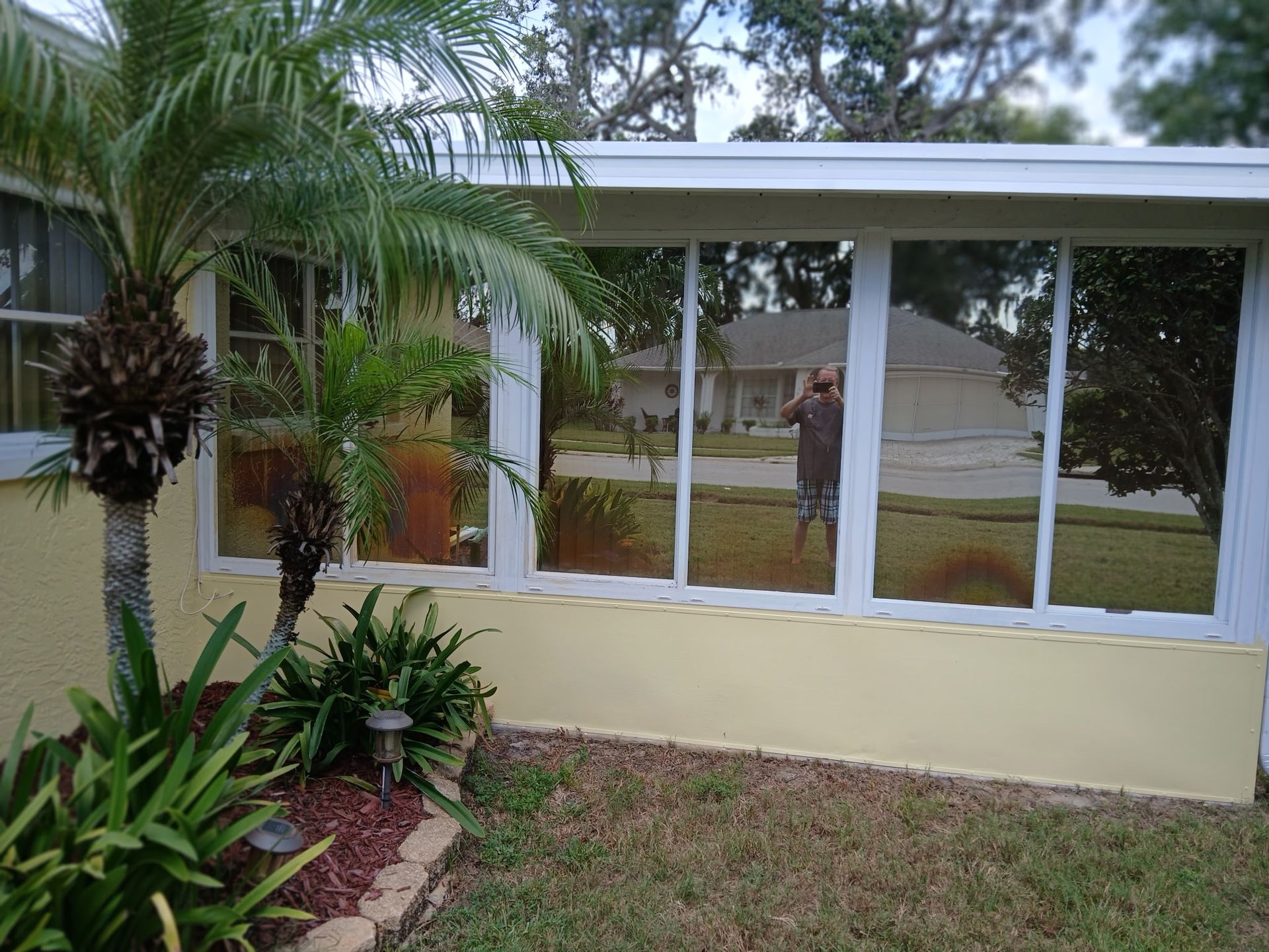 Single-story house with large windows, cream exterior, and tropical plants in the front yard.