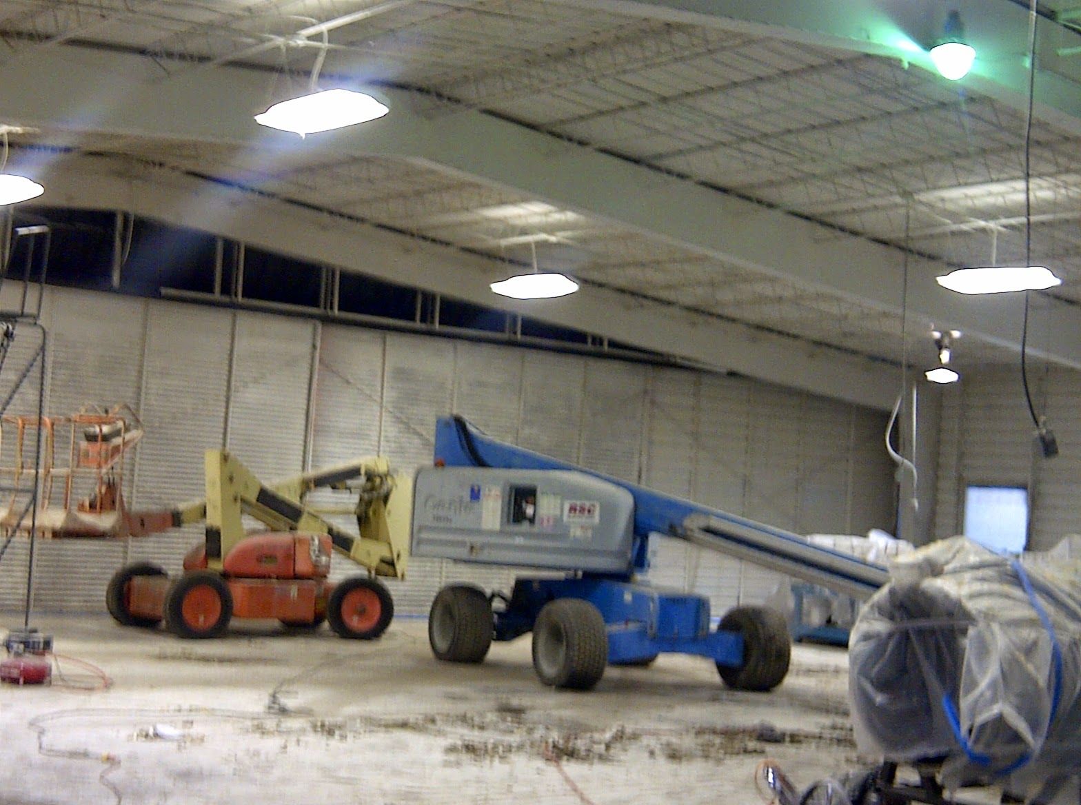 Warehouse with two cherry pickers, one orange and one blue, parked under a high ceiling.