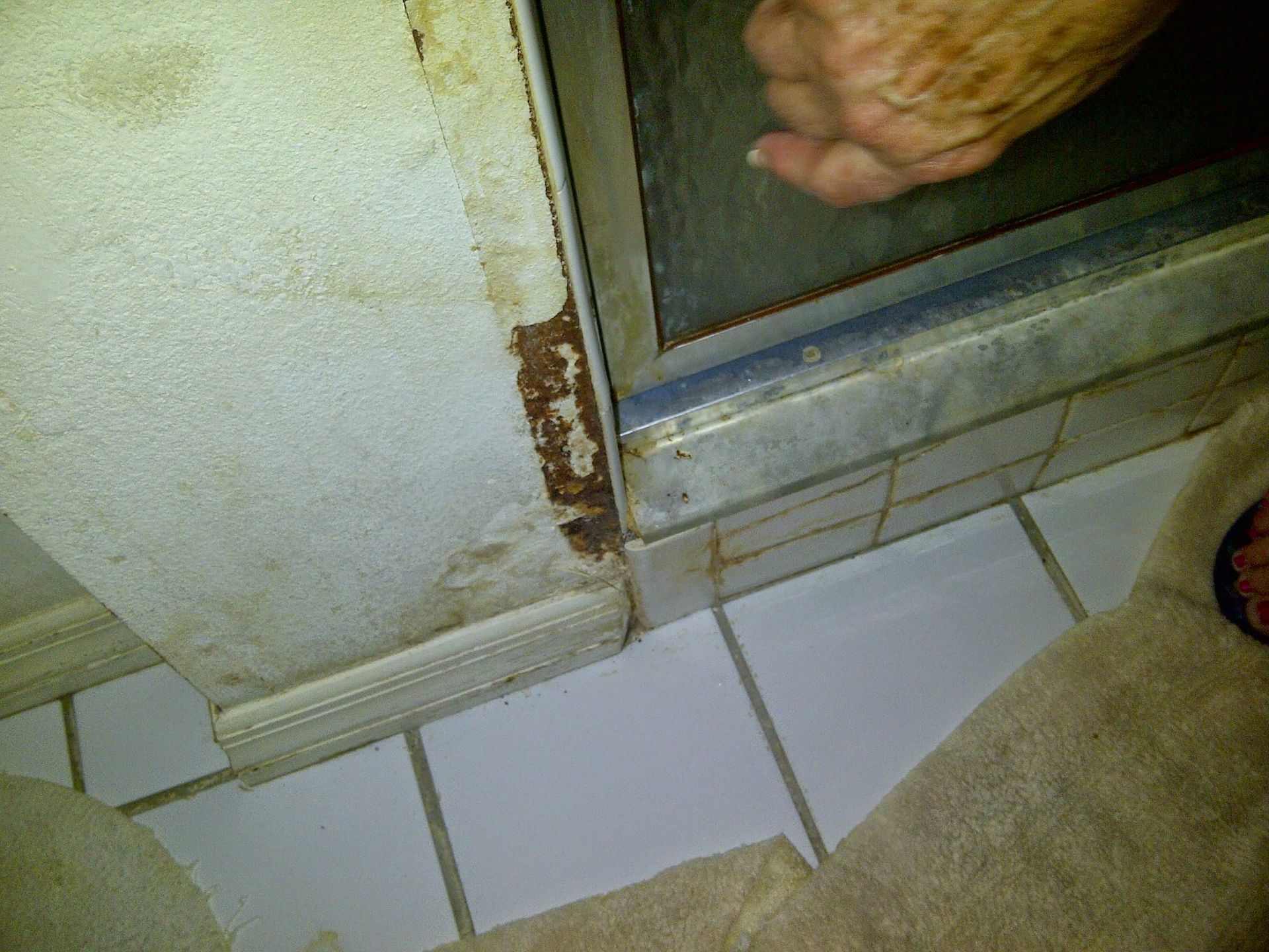 Rusty bathroom wall edge beside a shower door above white floor tiles, with a foot visible inside the shower.