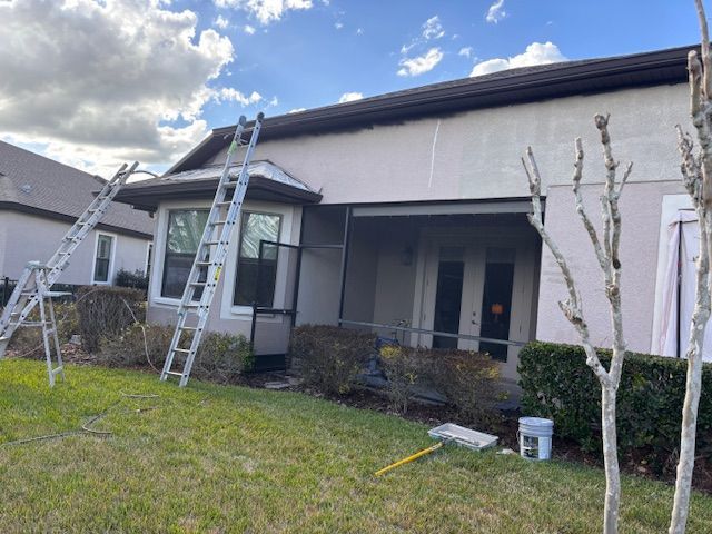 Two ladders lean against a house under repair, with a front porch, lawn, and partly cloudy sky.