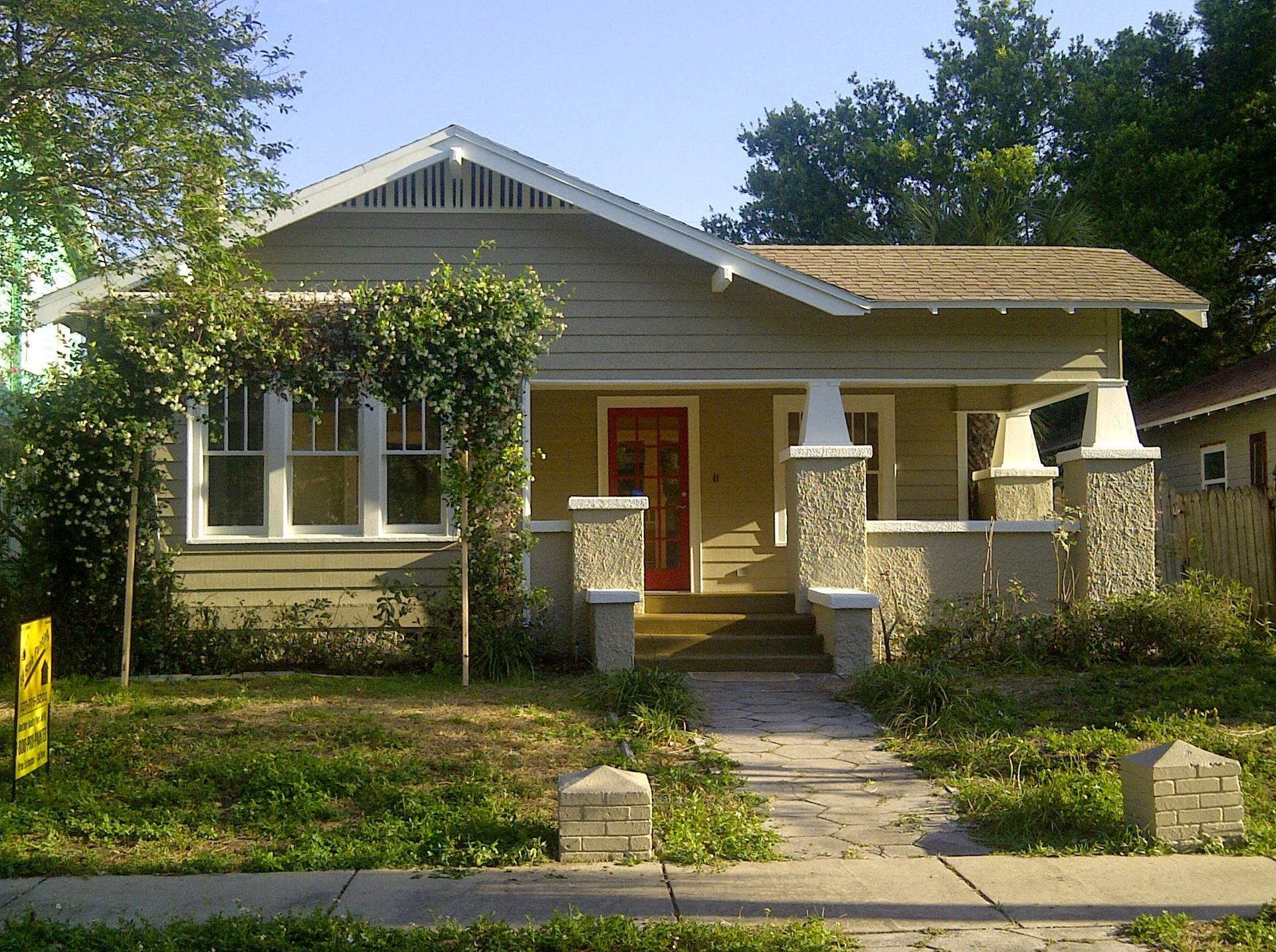 Small beige bungalow with a front porch, stone pillars, and a red front door behind a sidewalk.