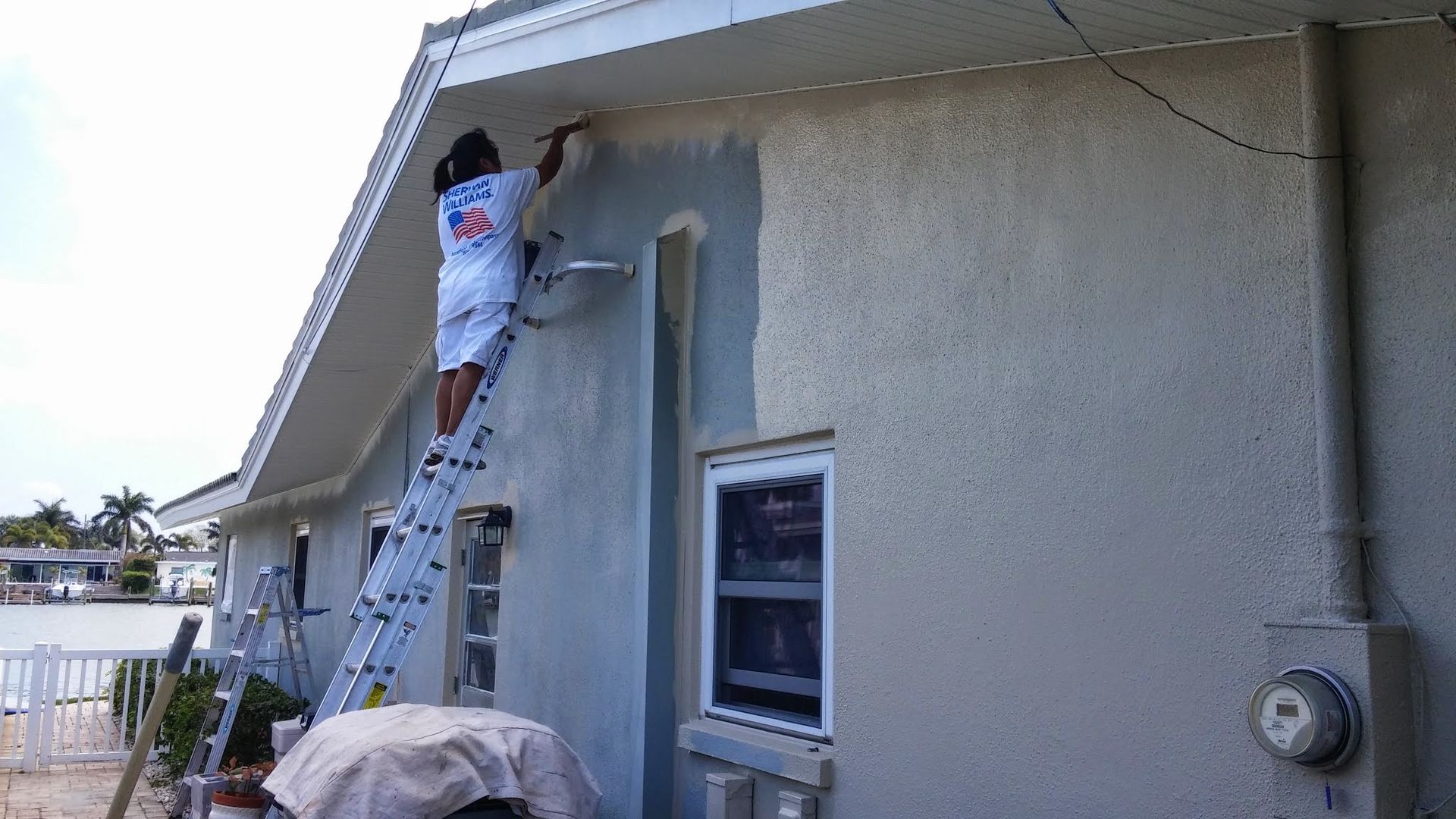 Person painting a stucco house exterior from a ladder near a window