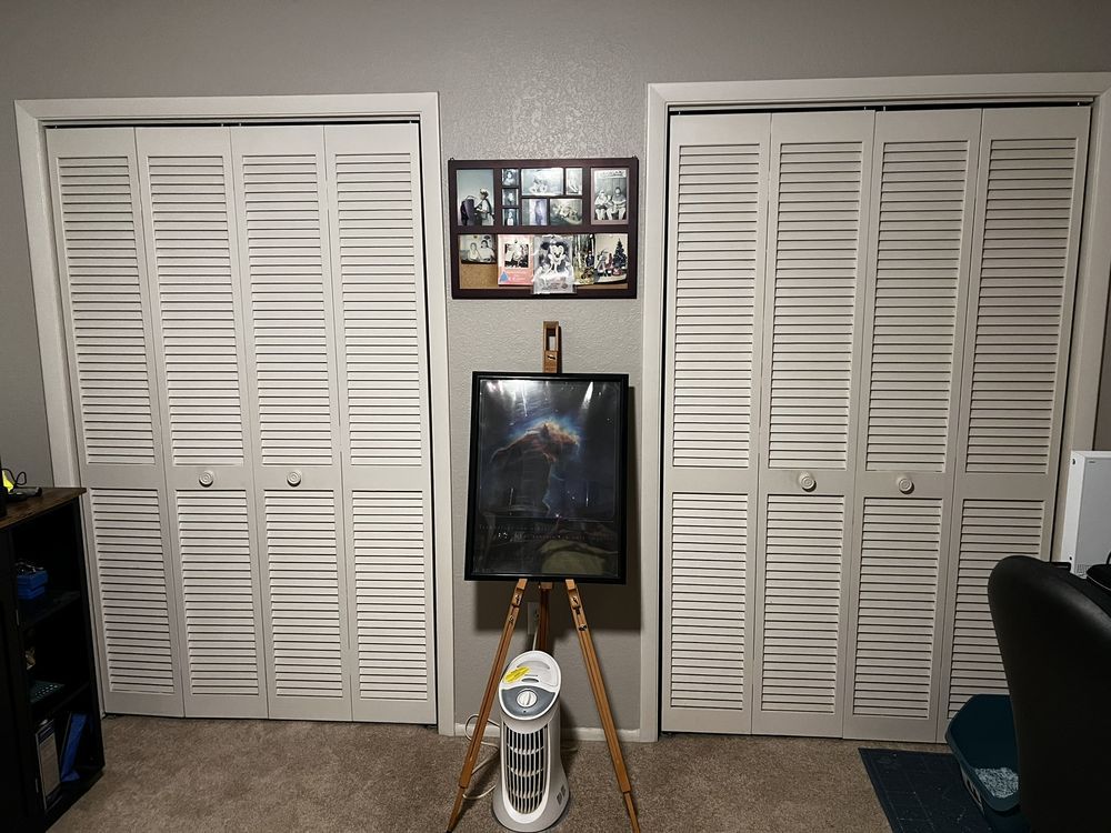 A room with white closet doors, framed wall photos, a painting on an easel, and a standing fan.