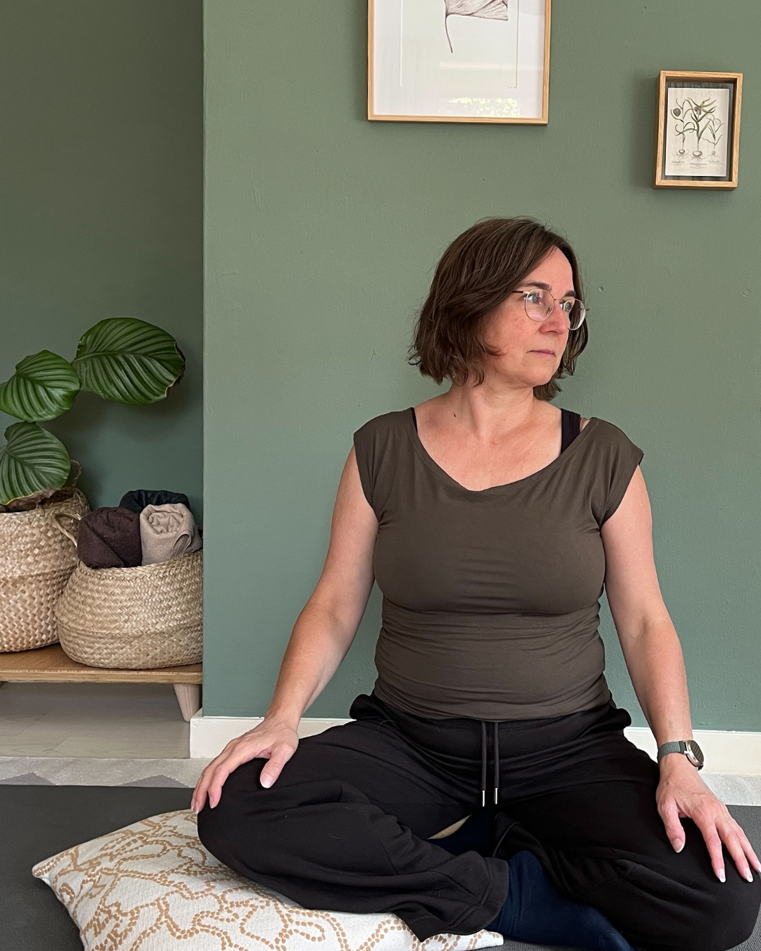 Astrid in zittende yogahouding, kijkt opzij; achtergrond: saliegroene muur.