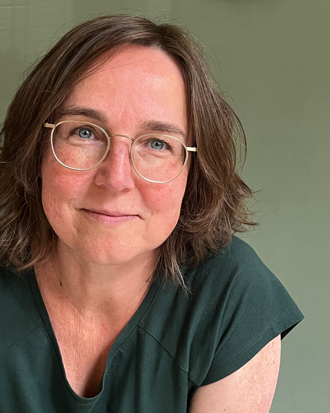 Astrid, vrouw met een bril en halflang bruin haar lacht, gekleed in een groen shirt, tegen een lichtgroene achtergrond.