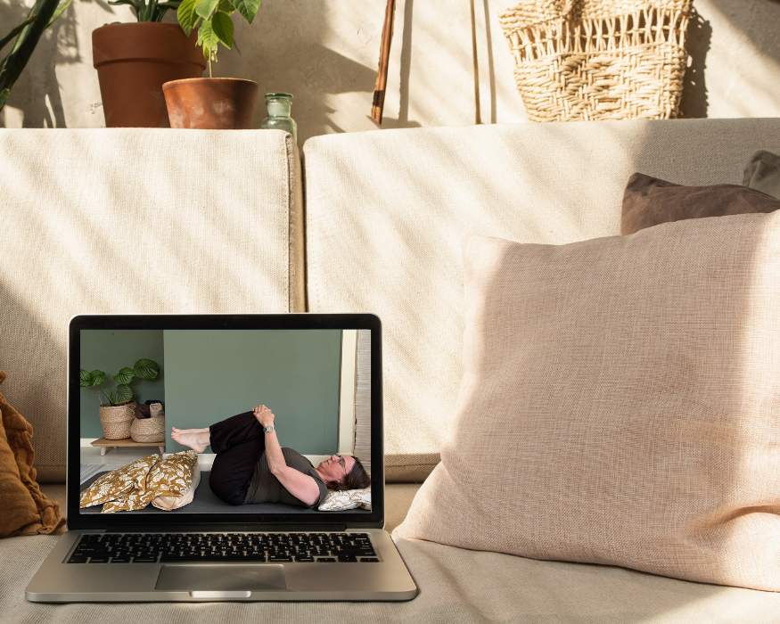 Laptop op de bank waarop een online yogales wordt weergegeven. Iemand ligt op de grond en houdt haar knieën vast. Zonnige binnenomgeving.