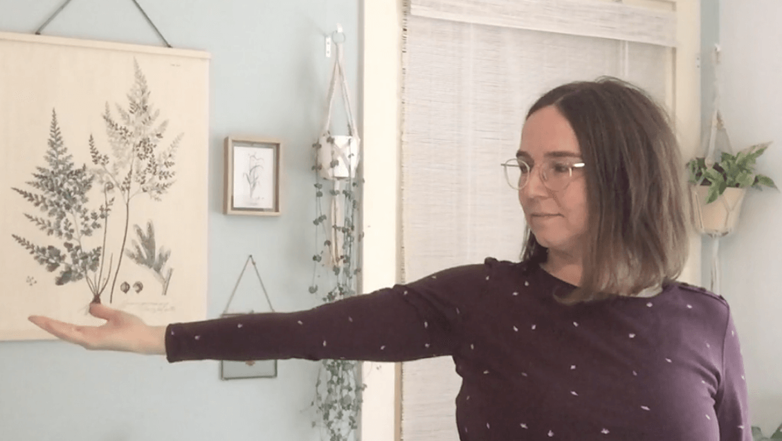 Astrid in een bordeauxrode blouse, strekt haar arm naar rechts uit met open handpalm, binnen, lichte achtergrond.