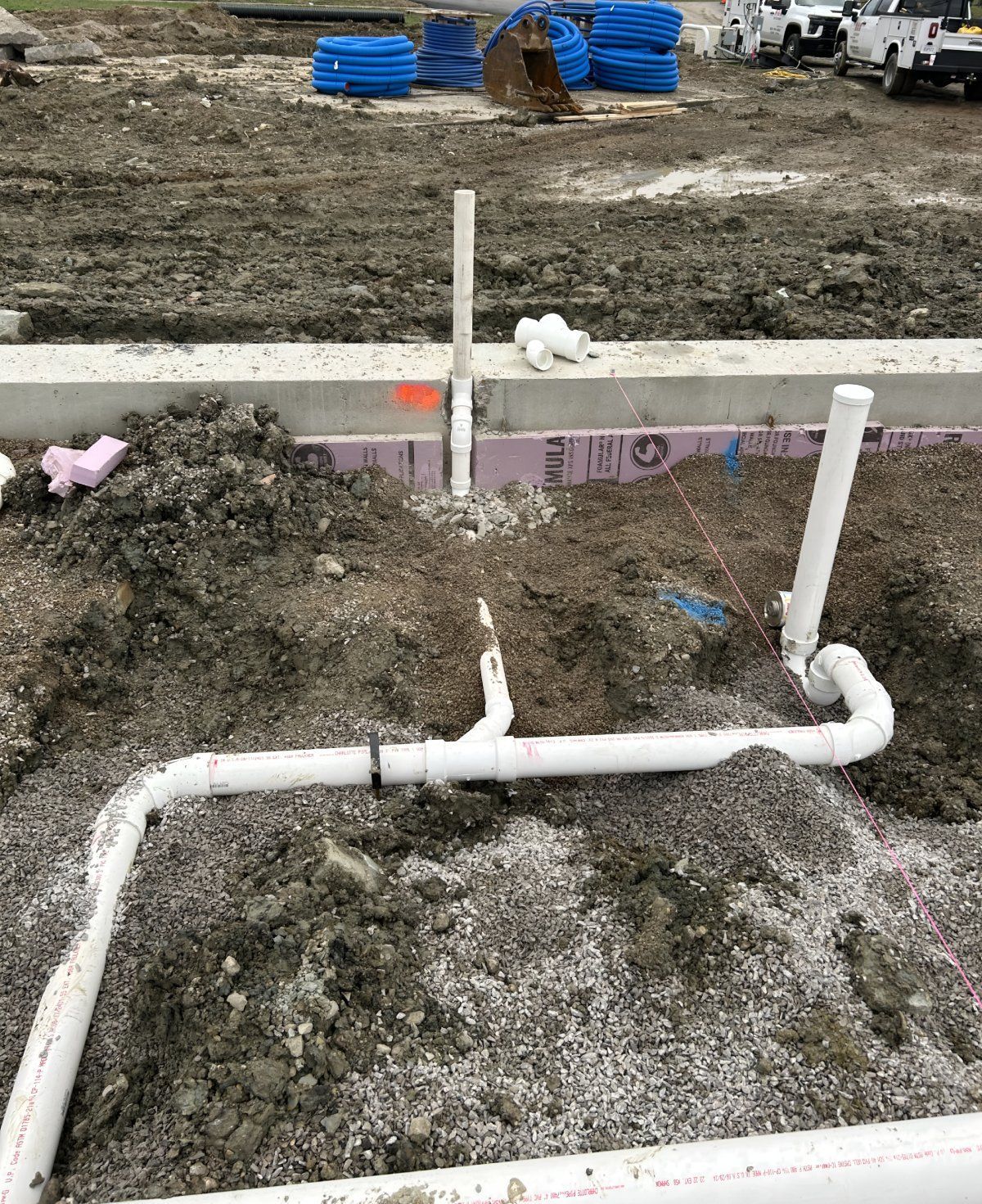 White PVC plumbing pipes emerge from the soil at a construction site, framed by a concrete foundation.