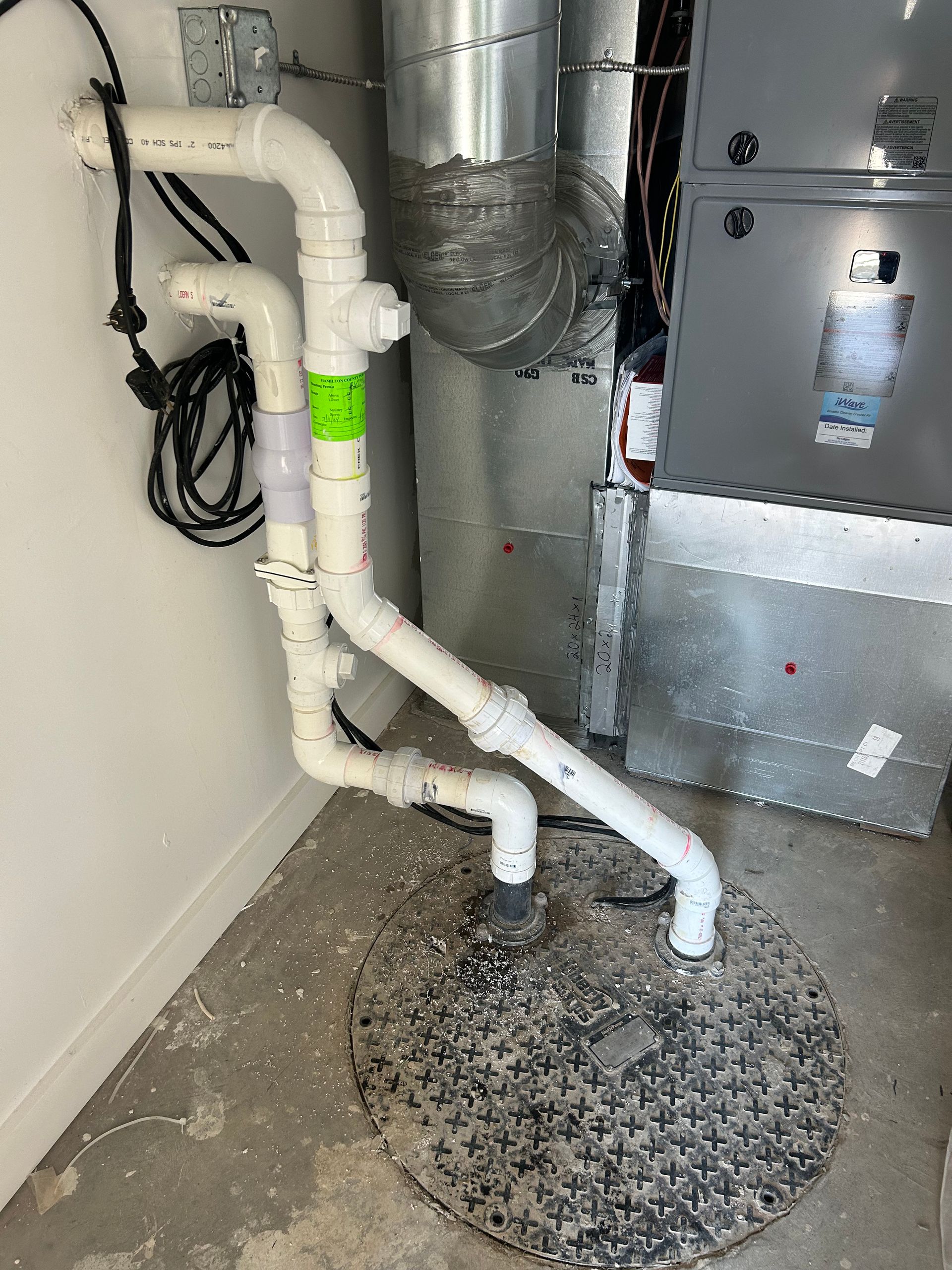 A sump pump and two white PVC pipes rising from a floor cover toward a wall and furnace in a utility area.