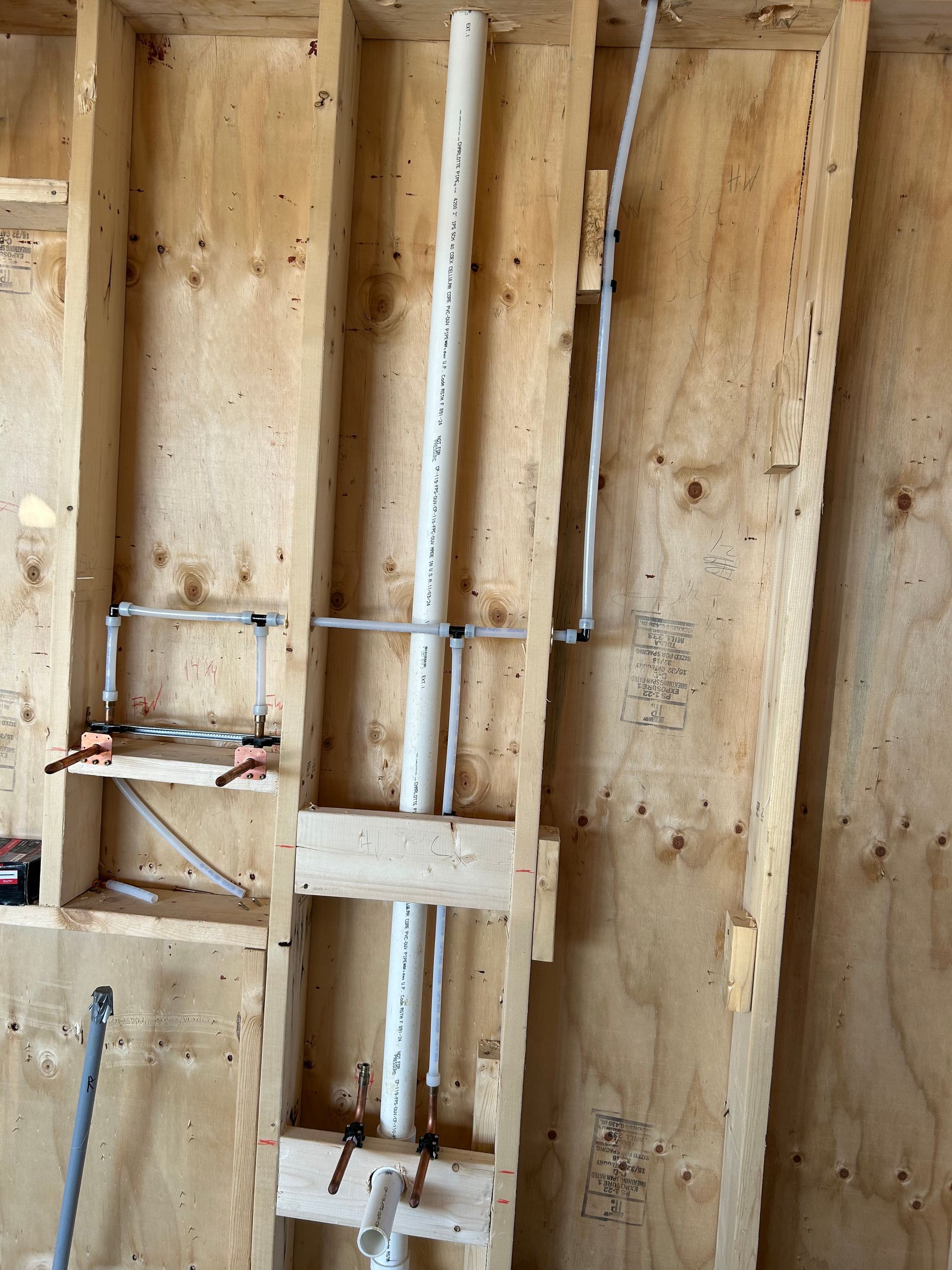 Plumbing pipes installed inside wooden wall studs during construction, featuring PVC drain and PEX supply lines.