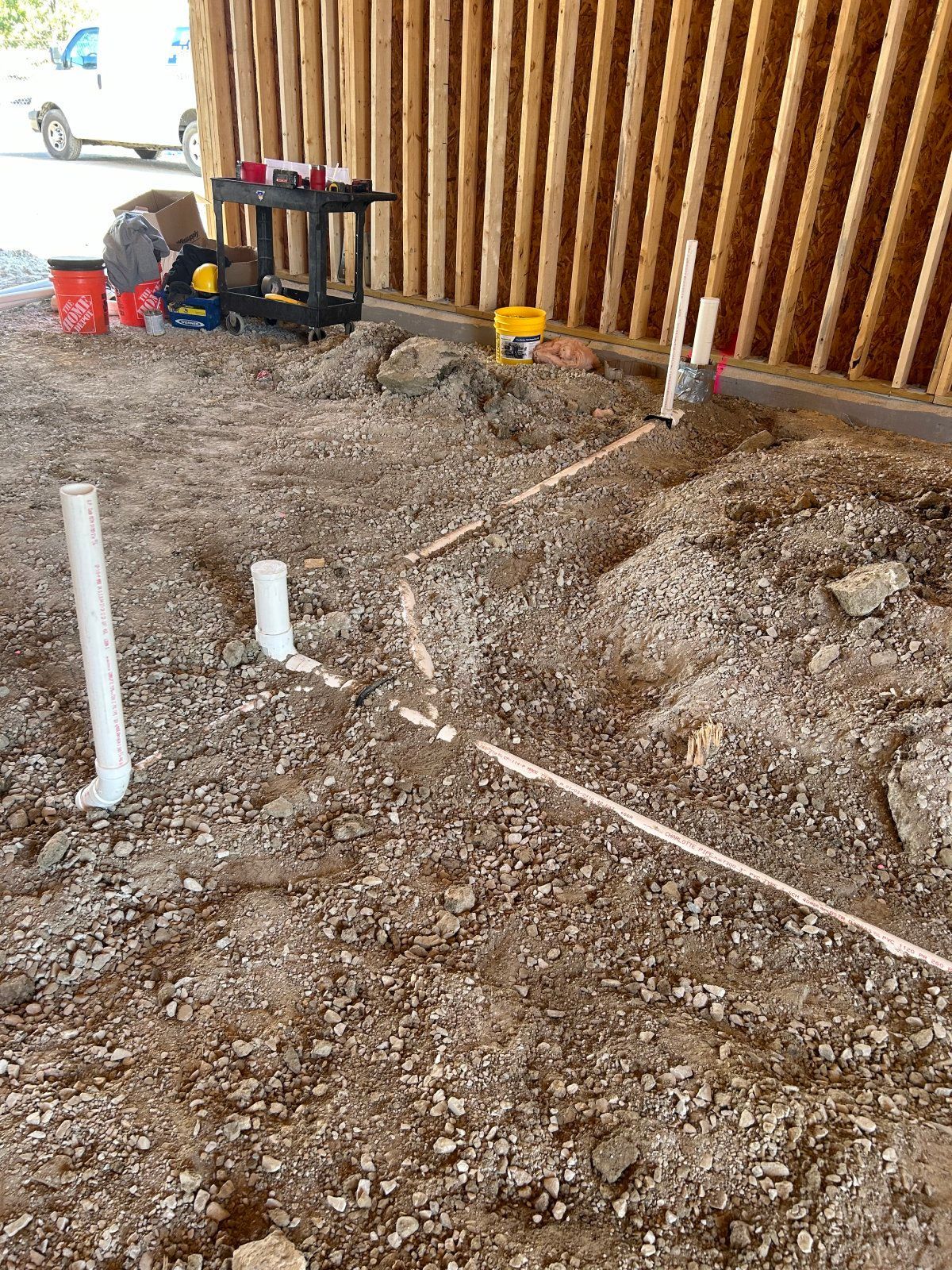 White PVC plumbing pipes emerge from gravel ground at a construction site near a wooden slat wall.