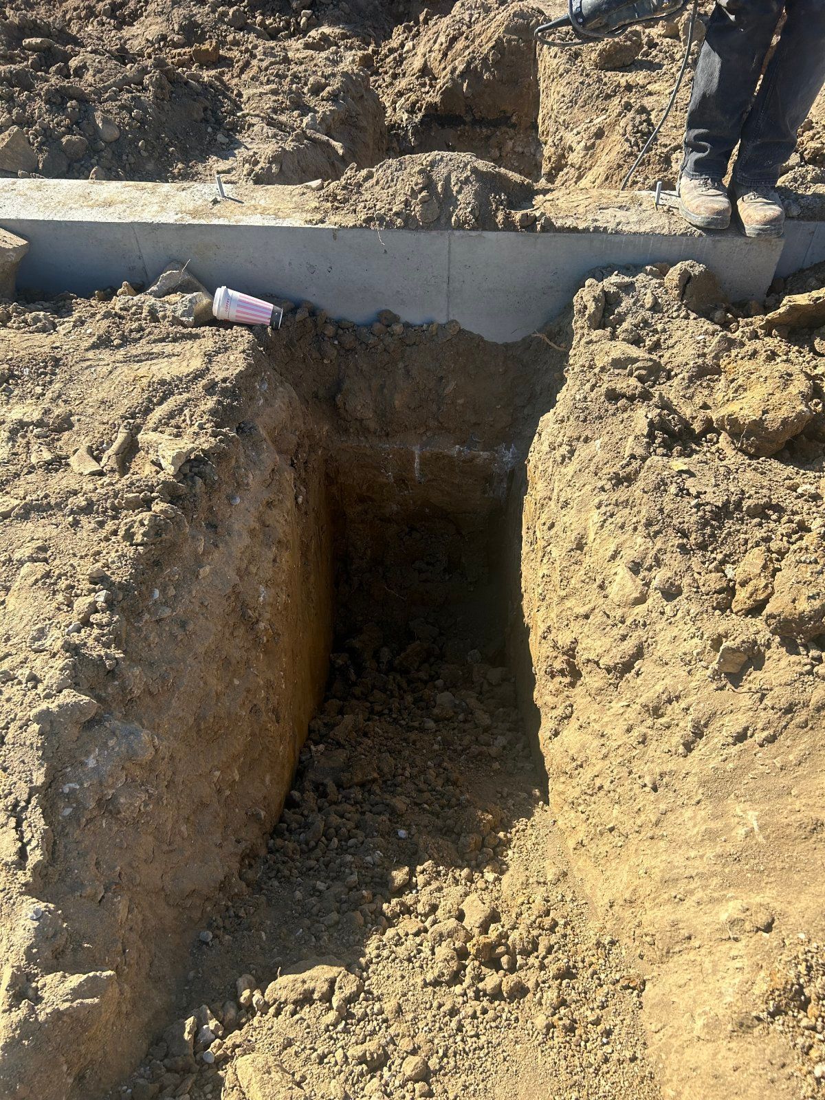 A deep, narrow trench dug into dirt leads up to a concrete foundation footer, with the legs of a worker visible nearby.