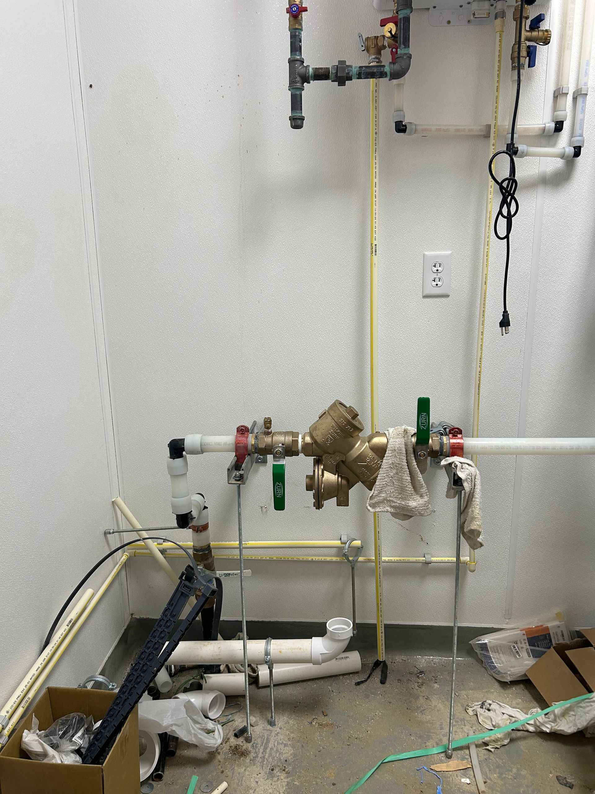 Exposed plumbing, valves, and white piping installed against a textured white wall in an unfinished utility room.