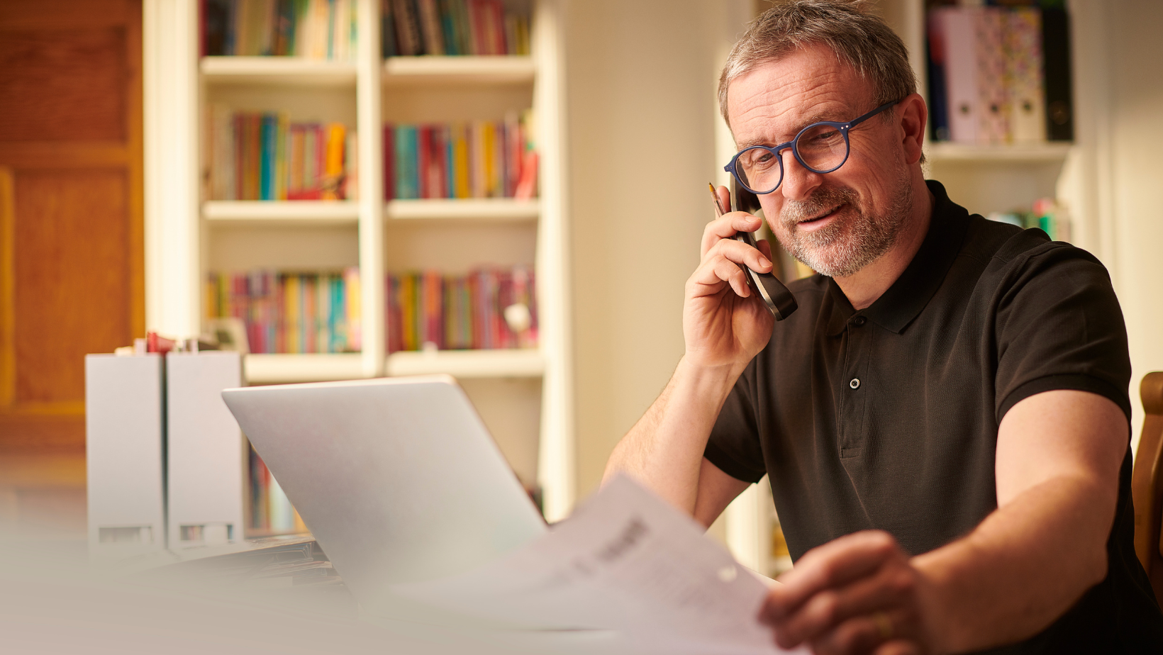 A man is sitting at a desk talking on a cell phone while looking at a piece of paper.