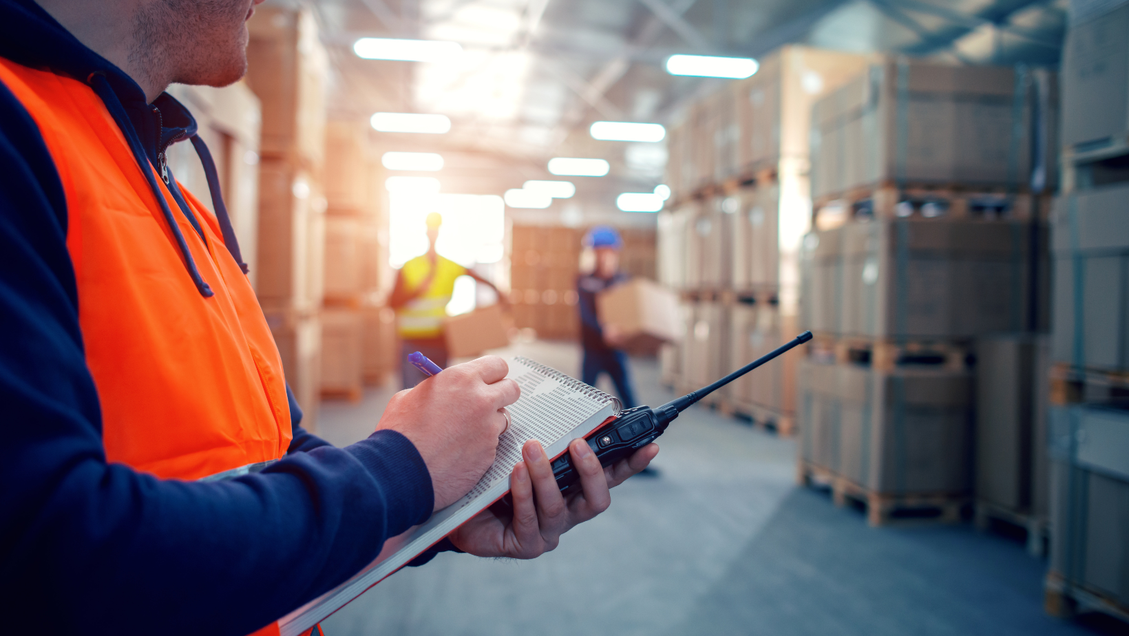 A man is holding a walkie talkie and a clipboard in a warehouse.