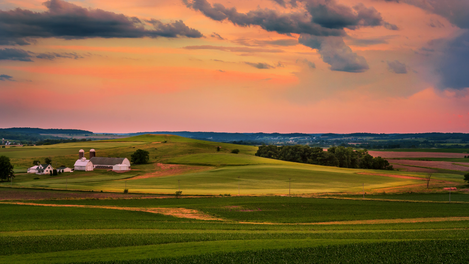 A farm in the middle of a field with a sunset in the background.