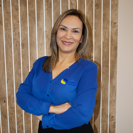 A woman in a blue shirt is standing with her arms crossed and smiling.
