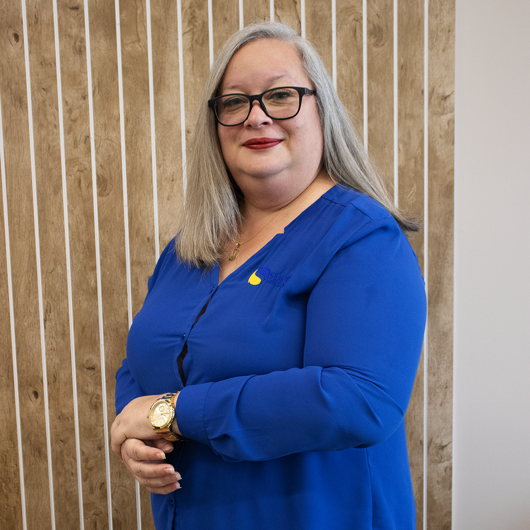 A woman wearing glasses and a blue shirt is standing in front of a wooden wall.