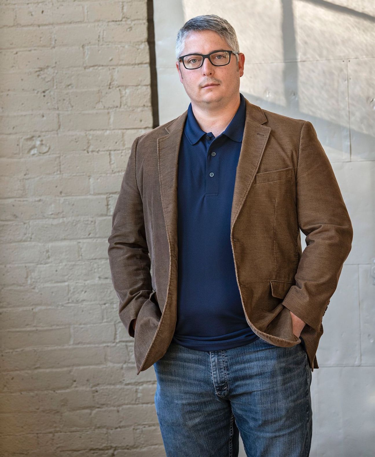 a man wearing glasses and a brown jacket is standing in front of a white brick wall .