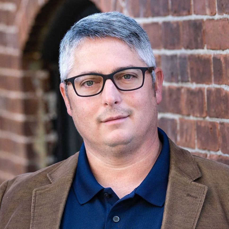 a man wearing glasses and a brown jacket is standing in front of a brick wall .