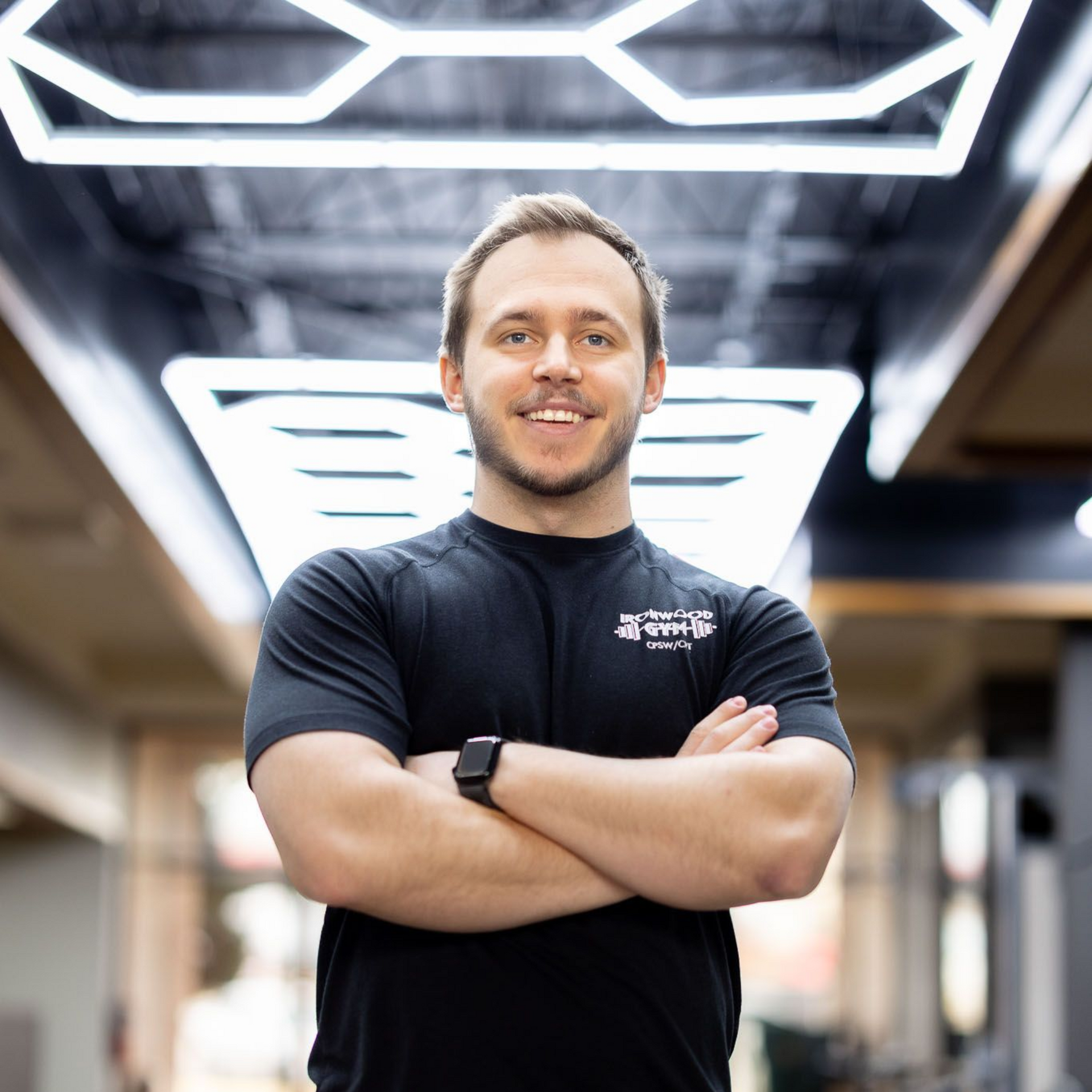 Smiling Trainer in a black shirt with arms crossed, standing under bright geometric ceiling lights indoors