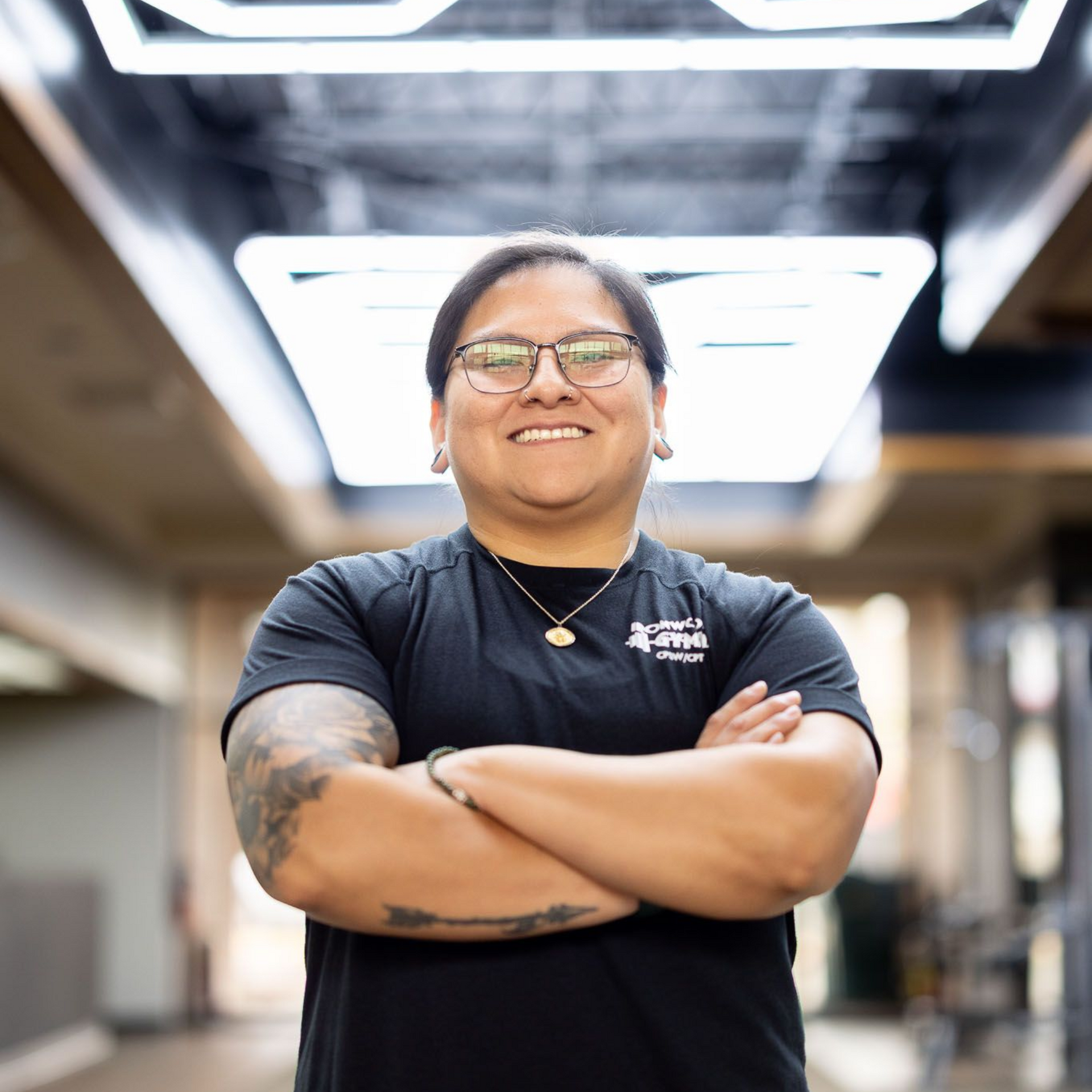 Smiling Trainer with crossed arms in a black shirt, standing under bright ceiling lights in Ironwood Gym