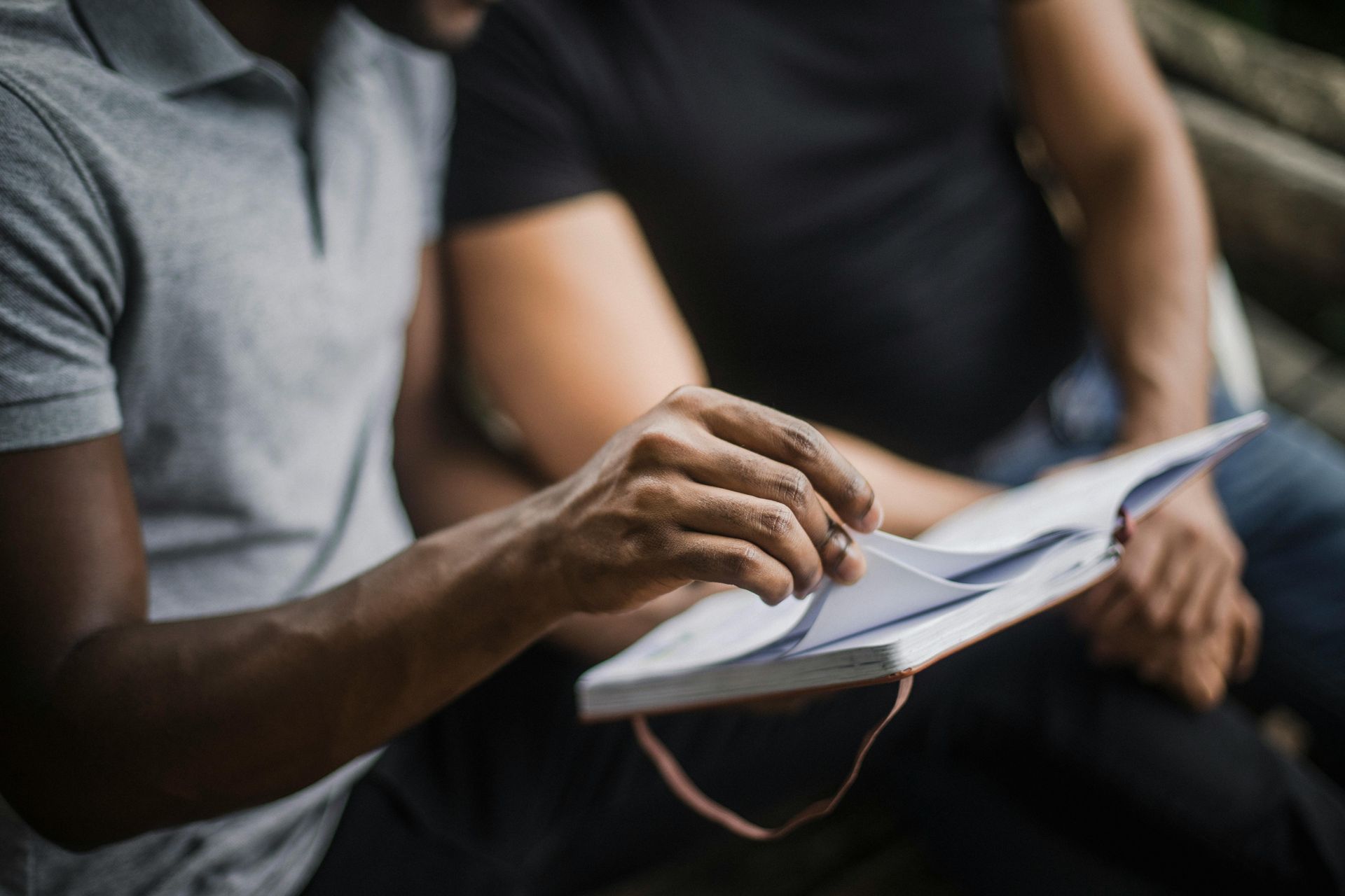Two people seated, one flipping through an open notebook or planner with a red ribbon bookmark.