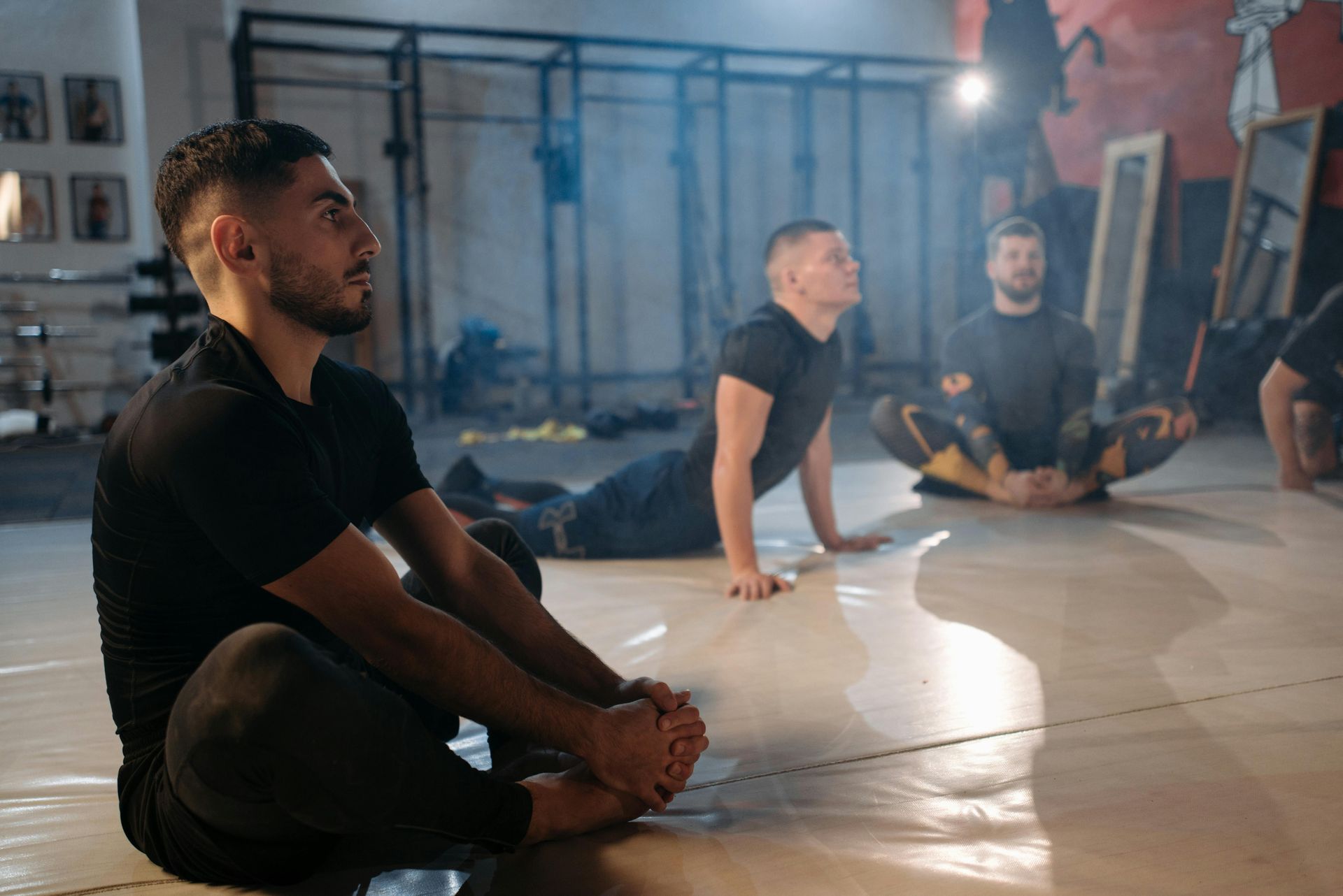 People stretching on mats in a gym studio, seated on the floor with a bright light in the background