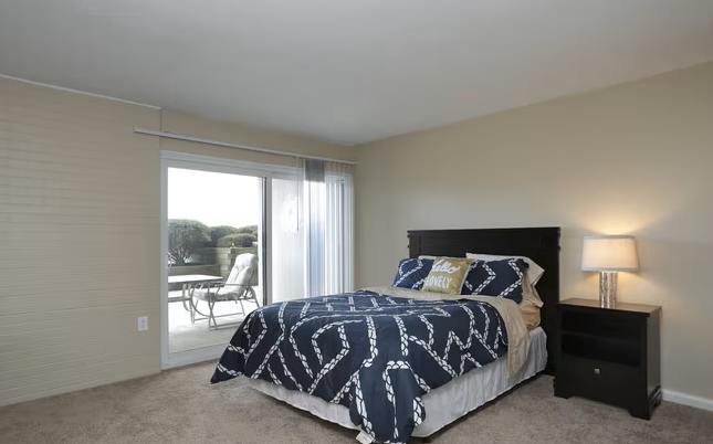 Bedroom with a neatly made blue-and-white bed, nightstand lamp, and sliding door to a balcony.