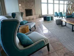 Modern lounge with teal armchairs, a stone fireplace, tiled floor, and large windows.