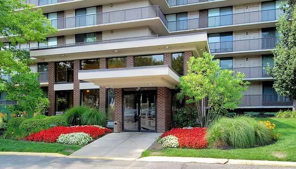 Apartment building entrance with glass doors, covered walkway, and red flower beds