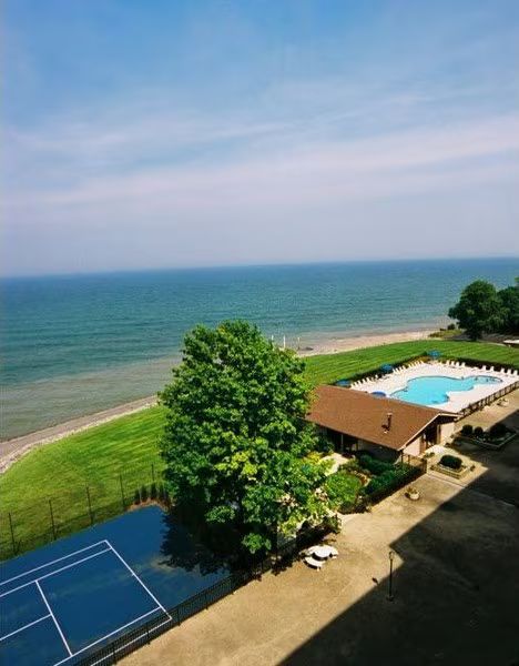 Lakefront resort with blue tennis court, pool, and trees beside a calm shoreline