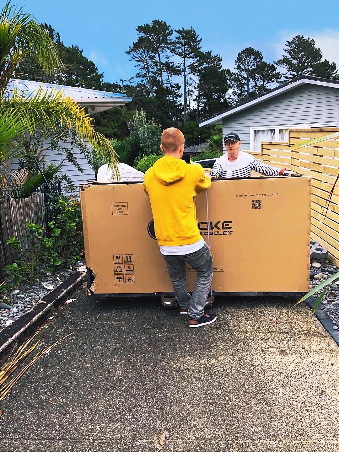A man in a yellow hoodie is standing next to a large cardboard box with an electric motorcycle inside it. Urban Classic New Zealand 2020