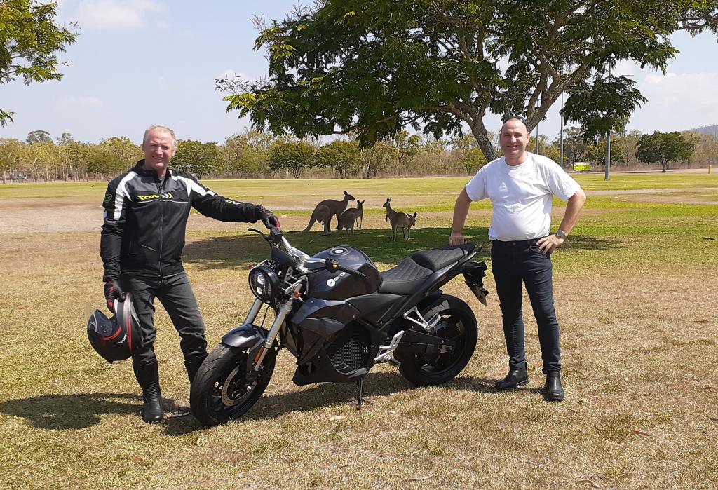 Two men are standing next to an Urban Classic electric motorcycle in a field with kangaroos in the background.