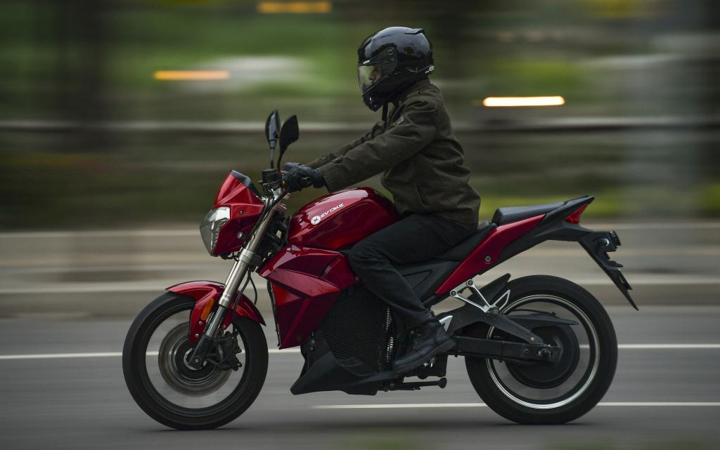 A man is riding a red urban-s, an electric motorcycle down a road.