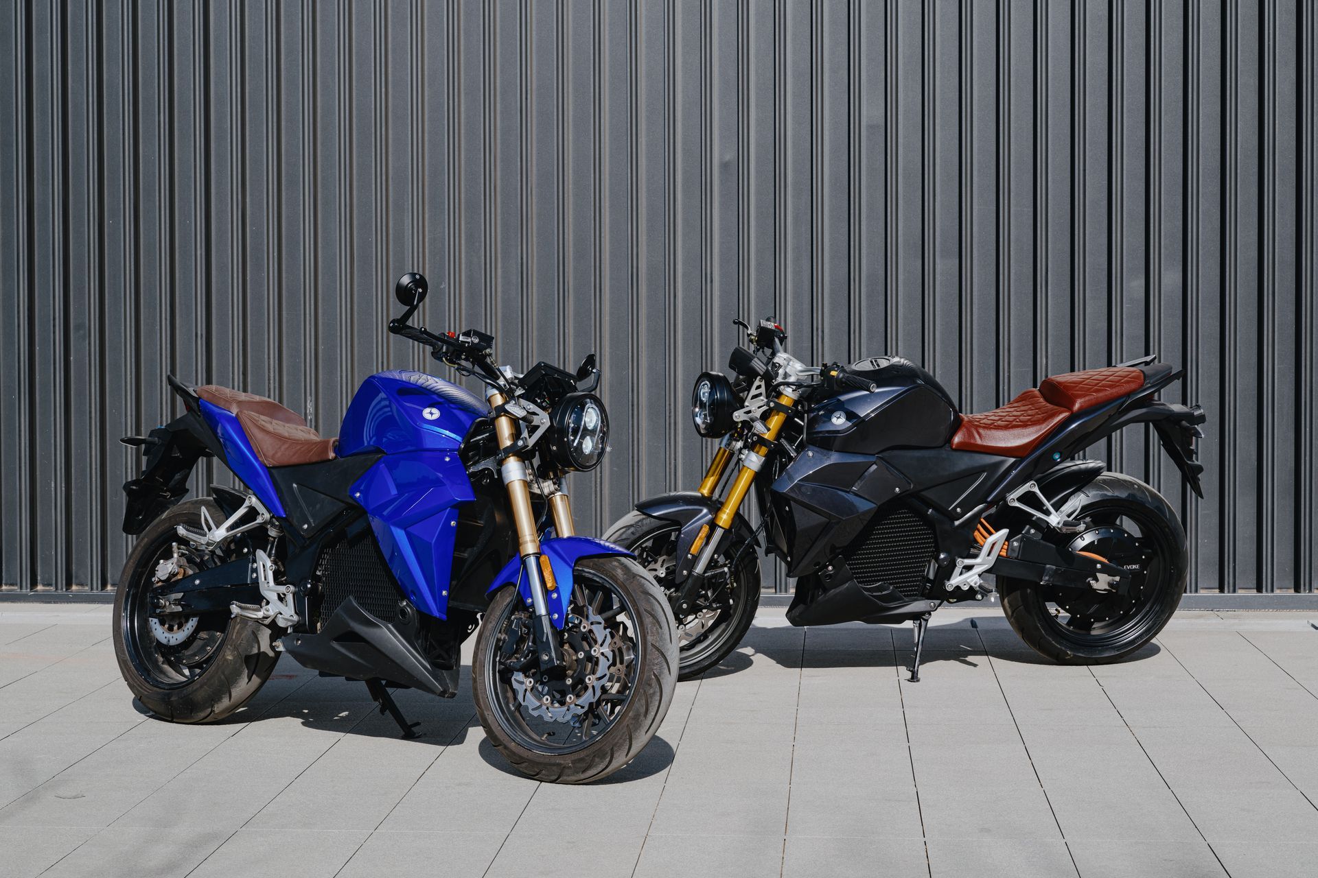Two electric motorcycles by EvokeMotorcycles are parked next to each other on a sidewalk in front of a building.