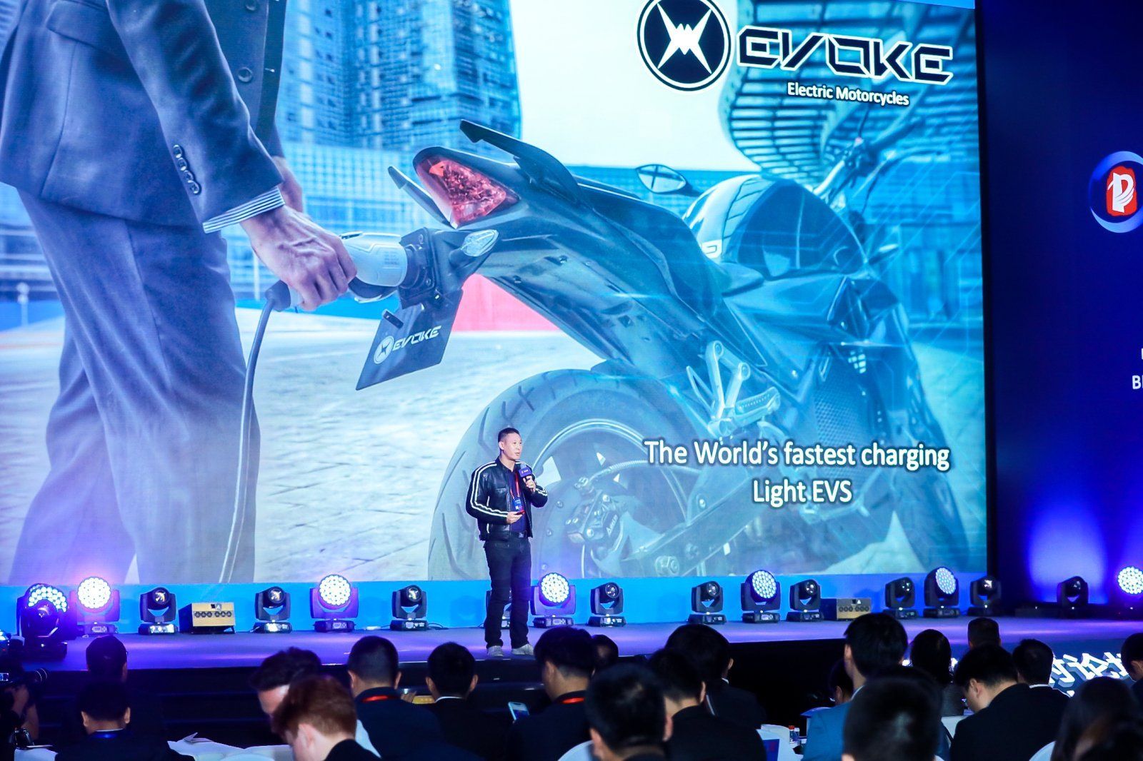 Nathan Siy is standing in front of a large screen with a picture of an electric motorcycle on it.