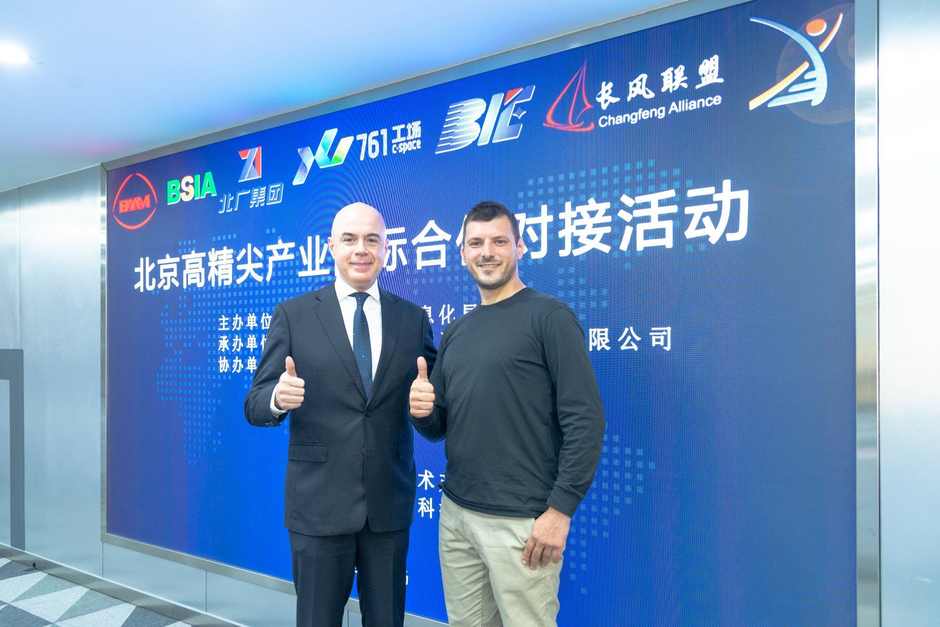 Two men are standing next to each other in front of a blue wall giving a thumbs up.