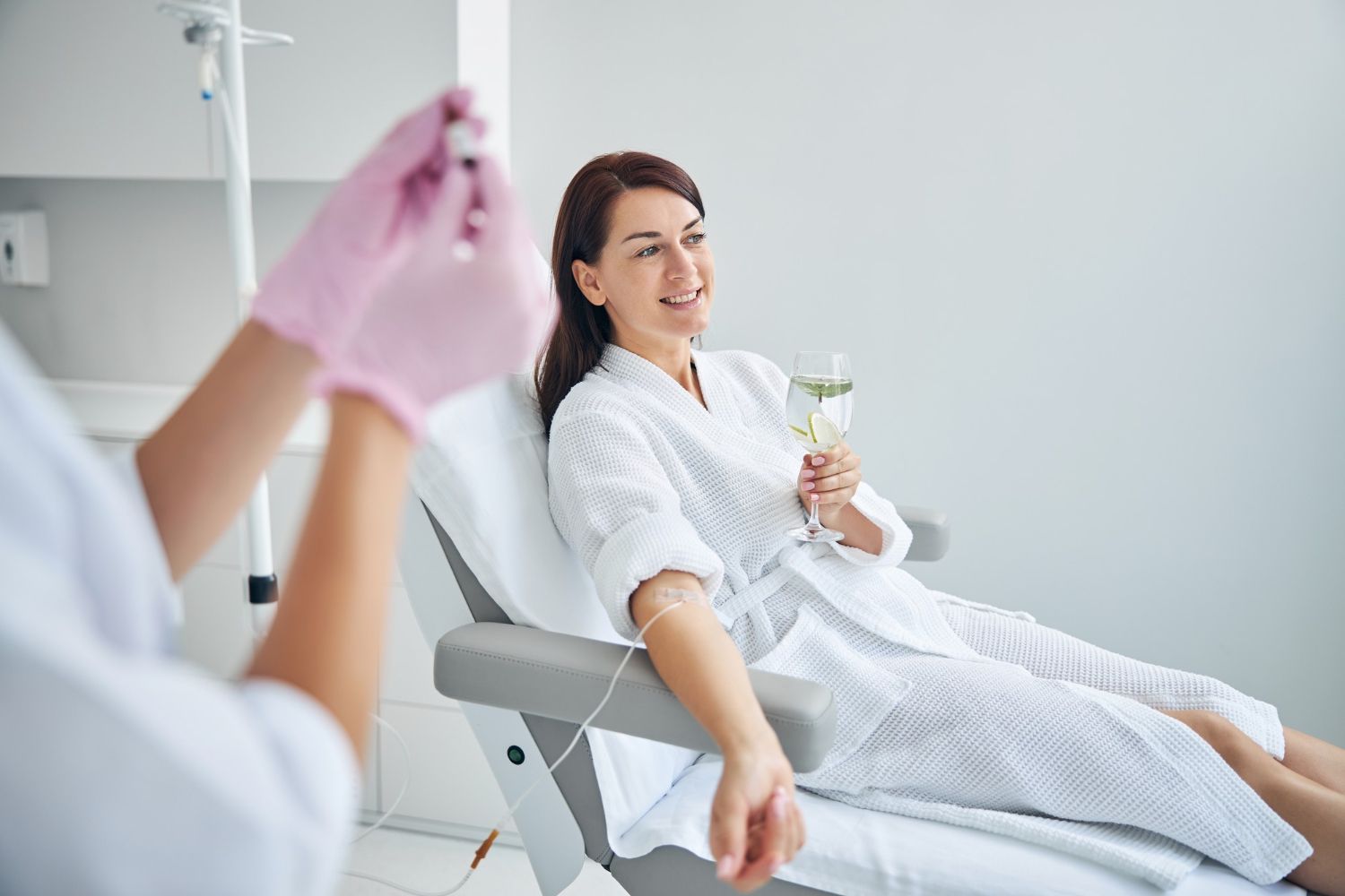 A woman is sitting in a hospital chair holding a glass of wine.
