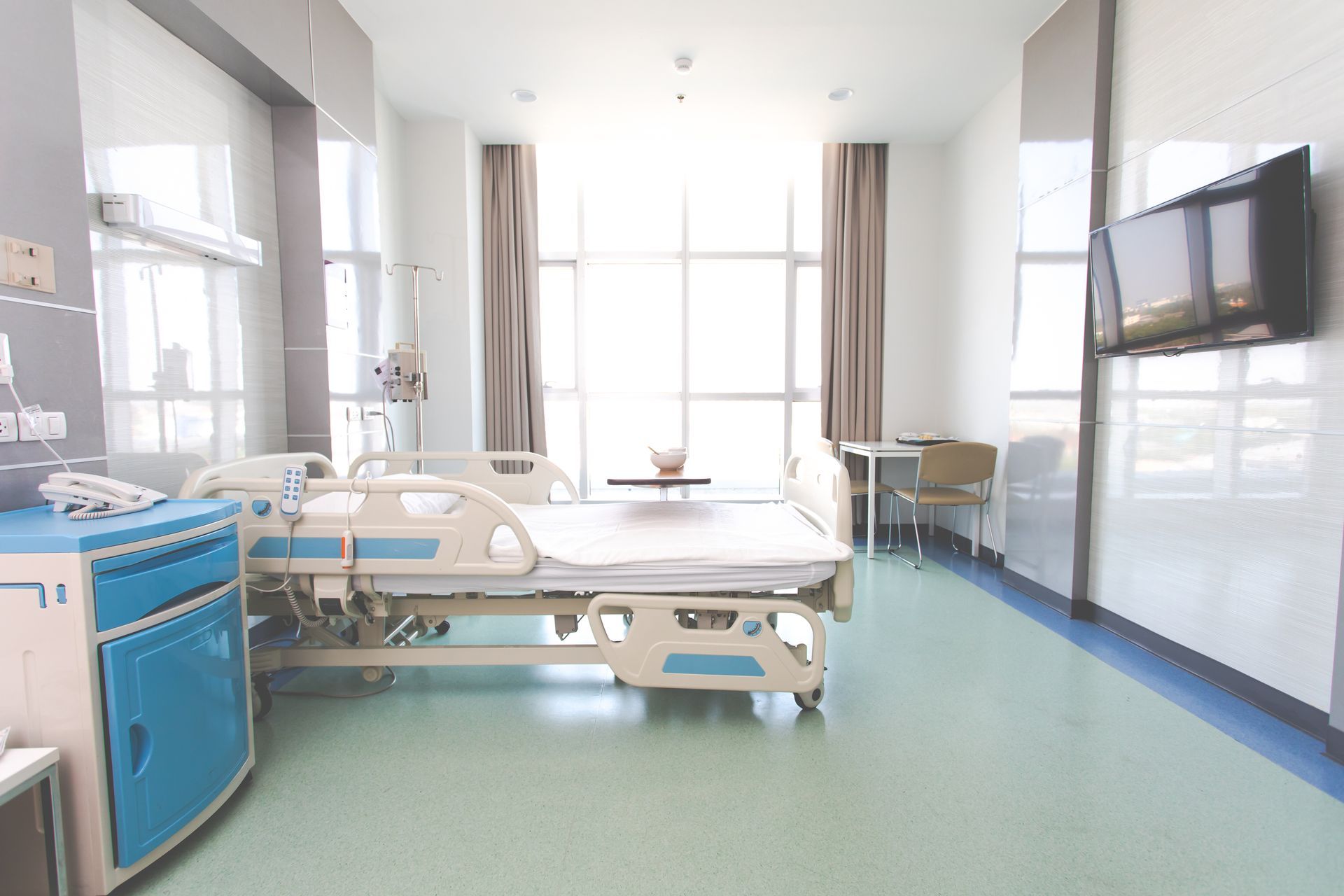 Bright hospital room with a bed, medical equipment, large windows, and a wall-mounted TV.