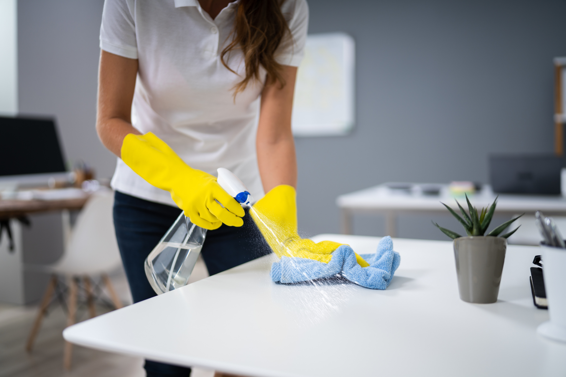 Person in yellow gloves cleaning a white desk with spray bottle and cloth in an office.