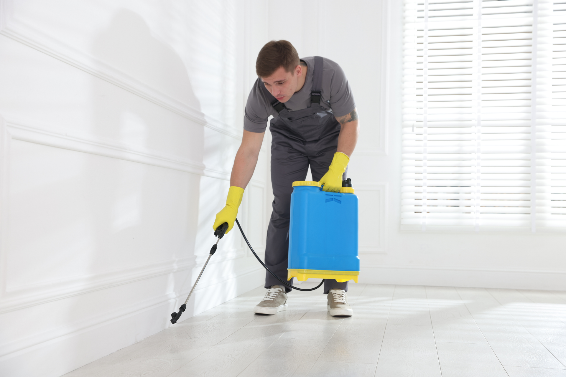 Person spraying a white floor with a blue backpack sprayer and black wand in a bright room