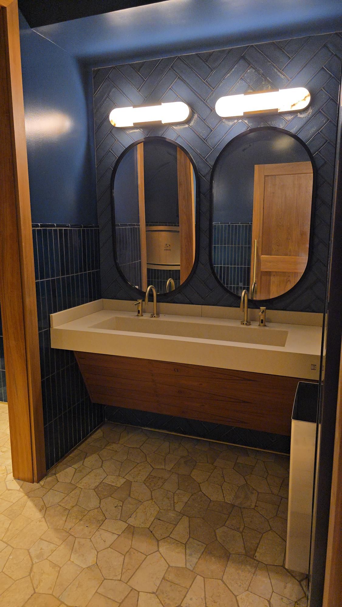Bathroom with dual sinks, oval mirrors, and blue tiled walls. The floor is patterned beige.
