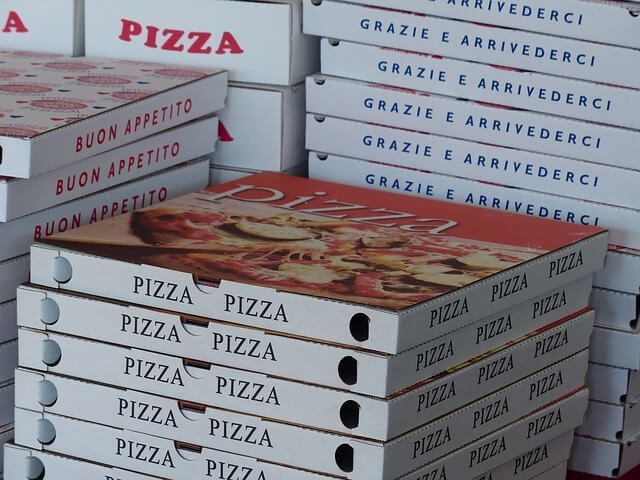 Scatole di pizza impilate su un tavolo, con stampate sopra le scritte “Pizza”, “Buon appetito” e “Grazie e arrivederci”.
