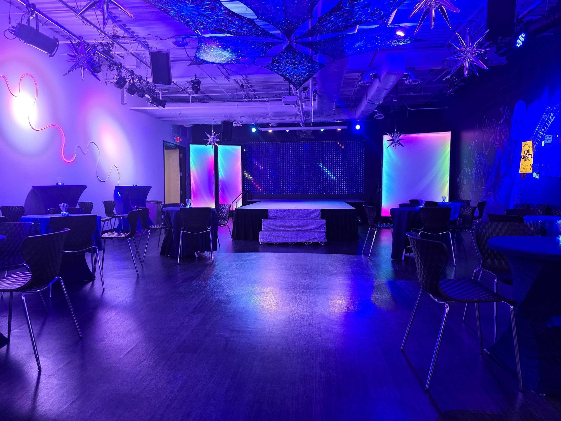 A performance space lit with blue and purple, featuring a stage, tables, chairs, and decorative lights.