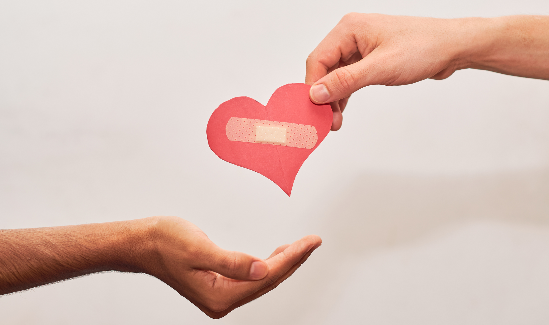 Hand giving a paper heart with a bandage to another open hand; white background.