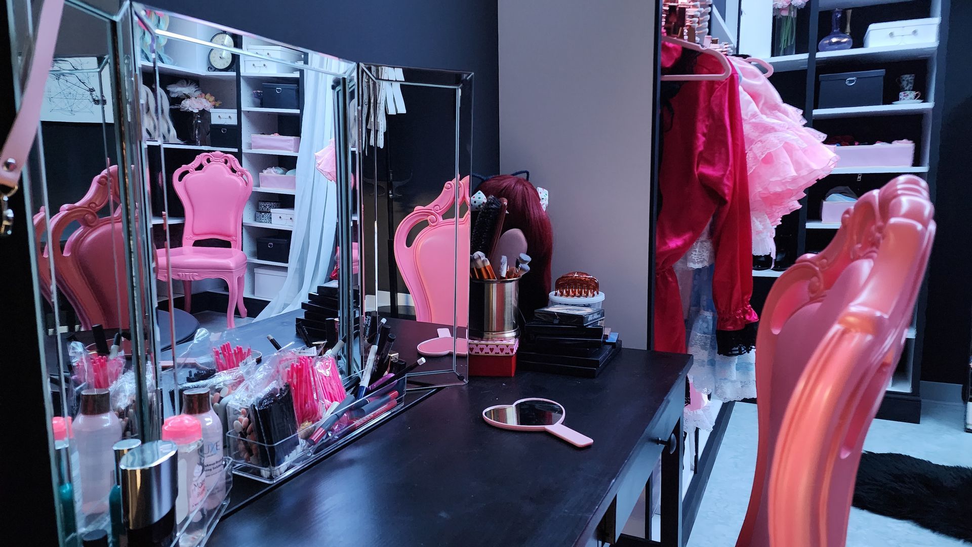 Pink and black vanity area with a desk, mirror, and makeup. Closet in the background with clothes.