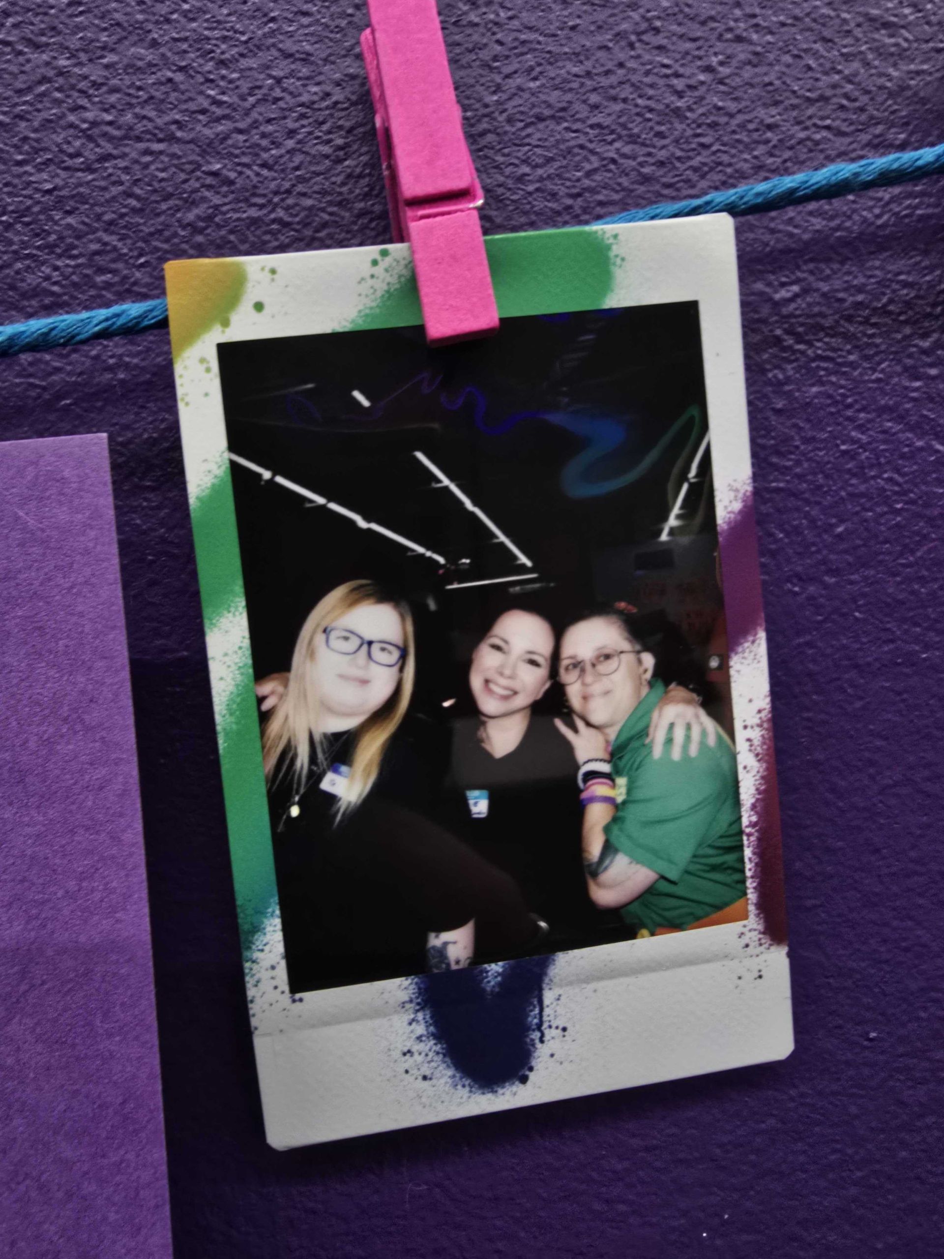 Three people pose for a photo. The photo is hung on a purple wall by a pink clothespin.