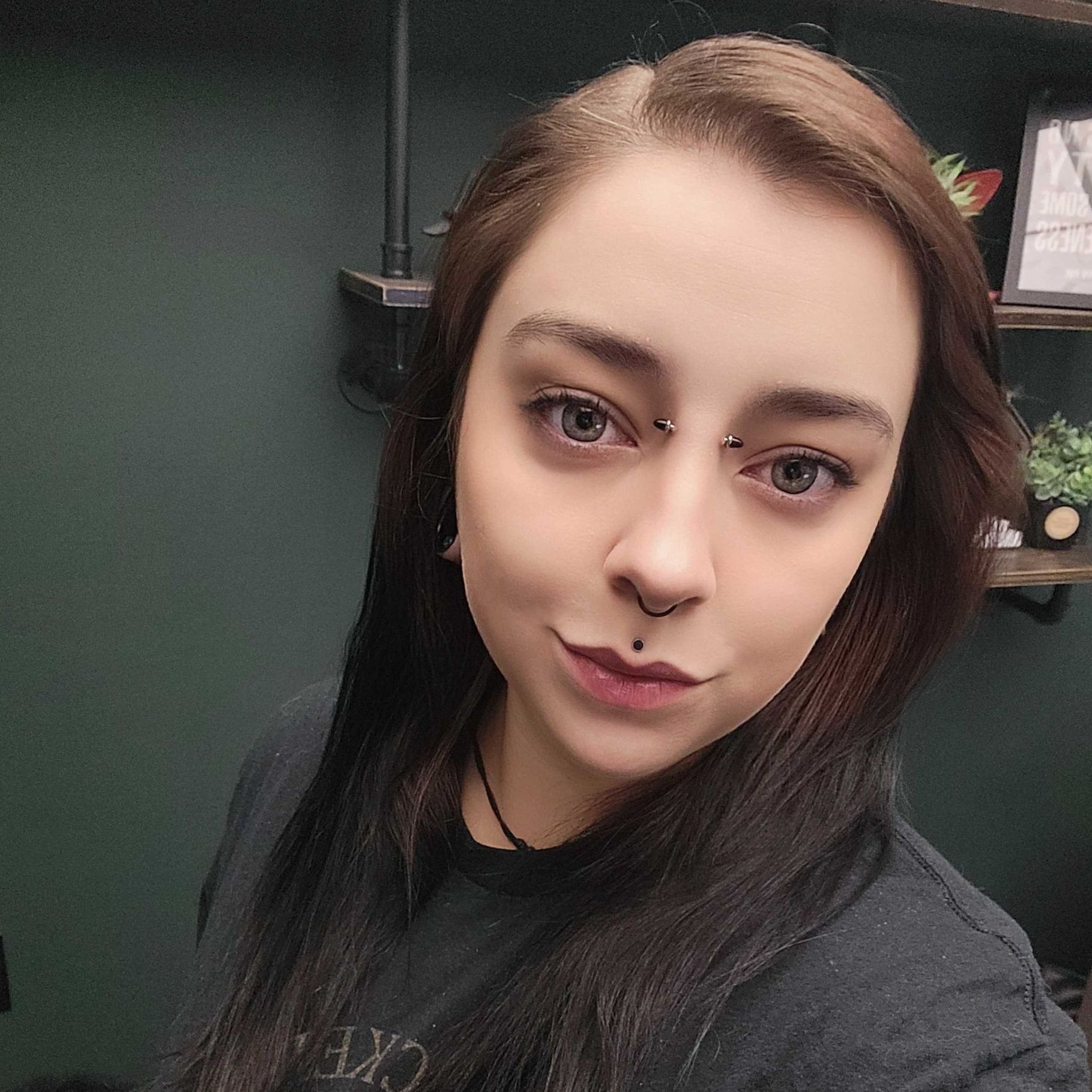 Daddy with multiple face piercings, looking at the camera. Dark hair, dark shirt, background with shelving.