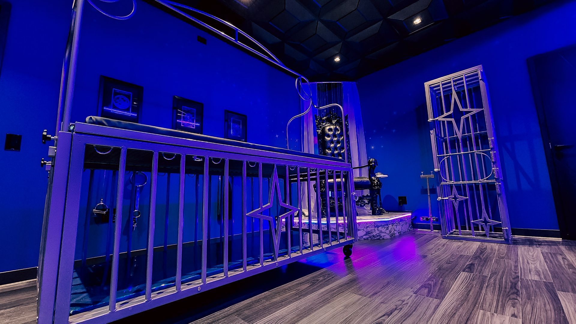 Blue-lit art installation with metal structures: a cage-like bed frame, a tower, and a central structure. Dark room.